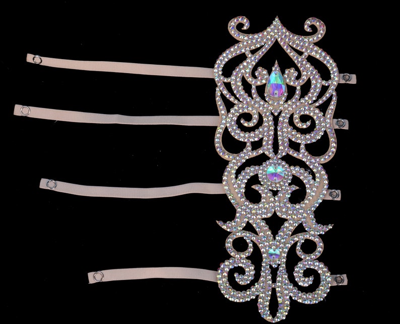 May include: A white, ornate, rhinestone-encrusted arm band with four adjustable straps. The arm band has a delicate, floral design with iridescent rhinestones.