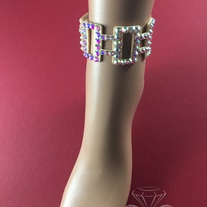 May include: Ankle bracelet with iridescent rhinestones set in a gold-tone metal. The bracelet features a rectangular design with multiple connected sections. The jewelry is displayed on a mannequin foot against a red background.