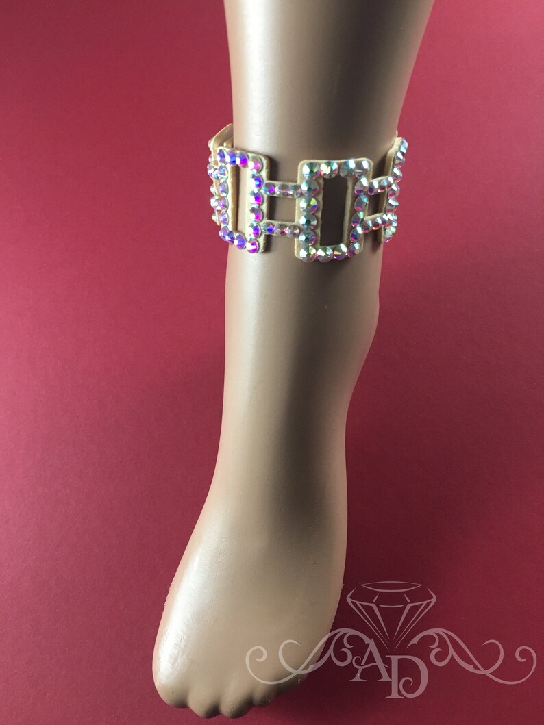 May include: Ankle bracelet with iridescent rhinestones set in a gold-tone metal. The bracelet features a rectangular design with multiple connected sections. The jewelry is displayed on a mannequin foot against a red background.