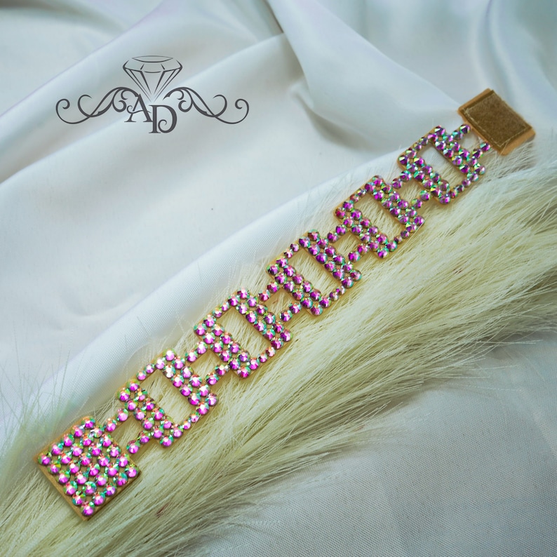 May include: A gold-toned bracelet with a geometric design, adorned with iridescent rhinestones. The bracelet features a rectangular clasp and is displayed on a white surface with a feathery accent.