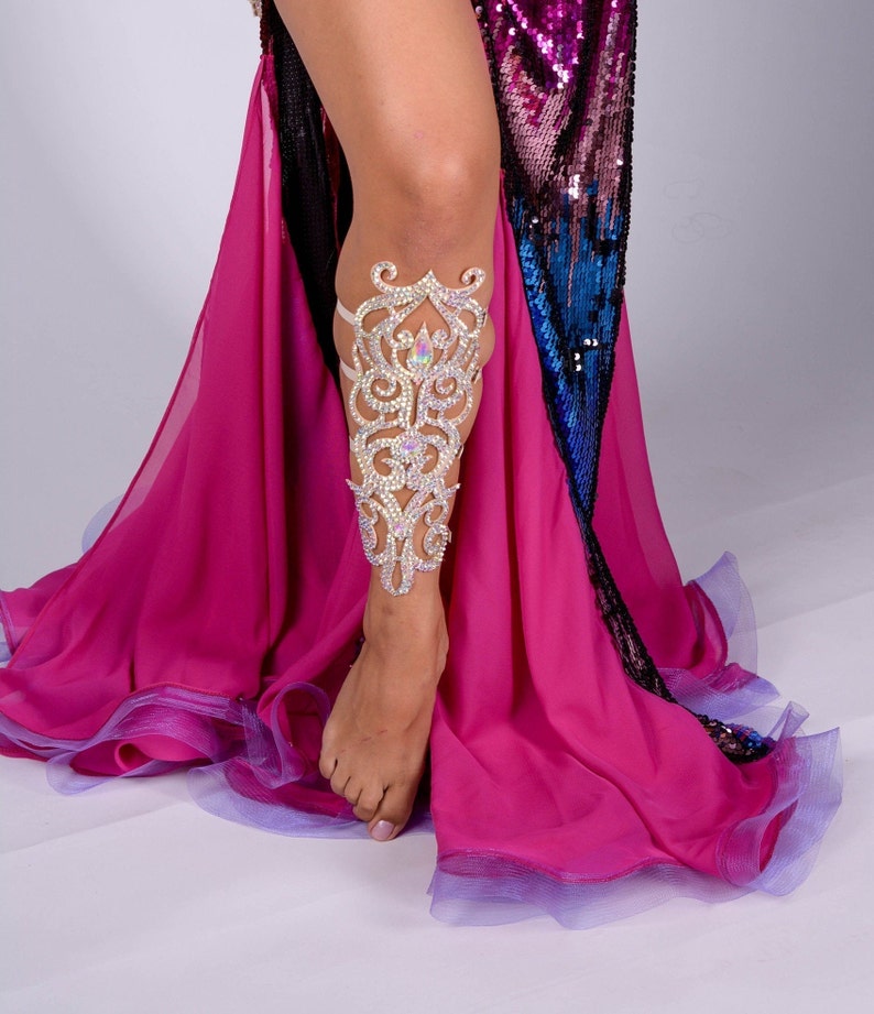 May include: A leg adorned with a sparkling silver leg cuff featuring an intricate floral design. The cuff is made of metal and has a delicate, feminine look. The leg is wearing a pink and blue sequined skirt.