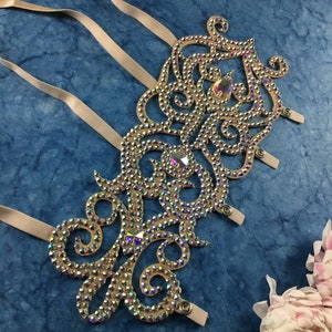 May include: A beige fabric arm band with a decorative design of swirls and loops covered in iridescent rhinestones. The arm band has three straps with snap closures.