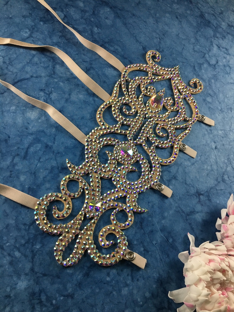 May include: A beige fabric arm band with a decorative design of swirls and loops covered in iridescent rhinestones. The arm band has three straps with snap closures.