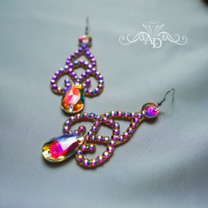 May include: A pair of dangle earrings with a gold base and iridescent crystals. The earrings have a teardrop-shaped crystal at the bottom.