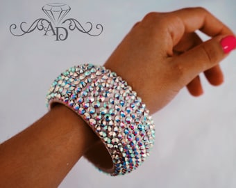 May include: A wide, iridescent rhinestone bracelet. The bracelet is covered in small, colourful rhinestones that catch the light. The bracelet is worn on a person's wrist, with a pink manicure visible. The bracelet has a light pink band.