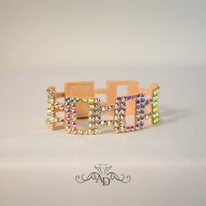 May include: A tan-colored bracelet with a geometric design. The bracelet features square-shaped links adorned with iridescent, multi-colored rhinestones. The bracelet is designed to be worn around the wrist.
