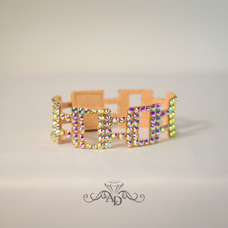 May include: A tan-colored bracelet with a geometric design. The bracelet features square-shaped links adorned with iridescent, multi-colored rhinestones. The bracelet is designed to be worn around the wrist.