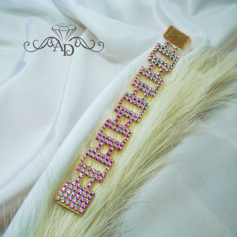 May include: A gold-toned bracelet with iridescent rhinestones arranged in a geometric pattern. The bracelet is set against a backdrop of white fabric and fluffy, light-colored decorative grass.