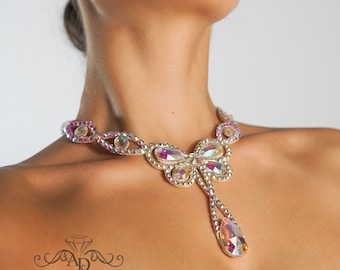 Puede incluir: Un collar de plata y de color iridiscente en forma de mariposa con un colgante en forma de lágrima. El collar está hecho de pedrería y tiene un diseño delicado.