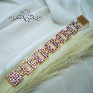 May include: A gold-toned bracelet with a geometric design, adorned with iridescent rhinestones. The bracelet features a series of square shapes connected by smaller links. The background is a white satin fabric and a feathery, light-colored plant.