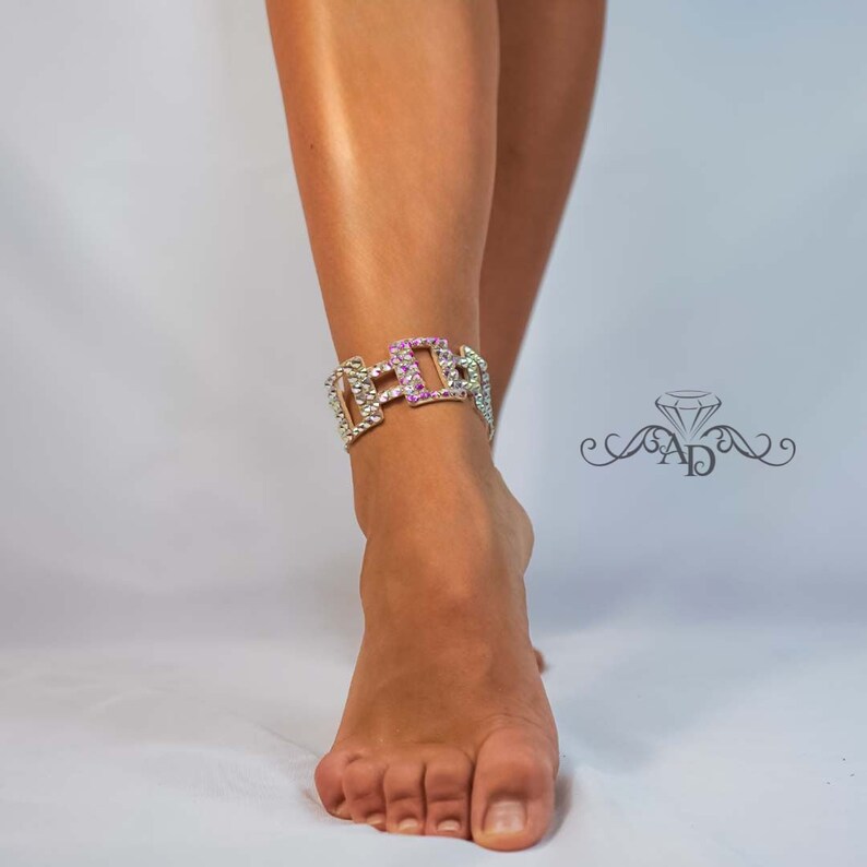 May include: A silver-toned anklet with a geometric design, adorned with sparkling rhinestones. The center section features pink stones, adding a pop of color. The anklet is worn on a foot against a white background.