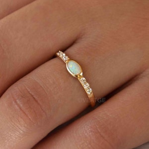 May include: A gold ring featuring an oval opal center stone, flanked by small, clear accent stones. The ring is set on a delicate gold band. The hand wearing the ring is resting on a white fabric. The brand name "VEATGE" is visible.