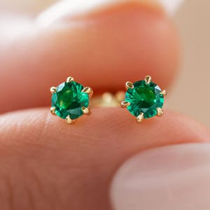 May include: A pair of gold stud earrings, each featuring a vibrant green gemstone. The gemstones are round-cut and set in a six-prong setting. The earrings are displayed against a soft, blurred background.