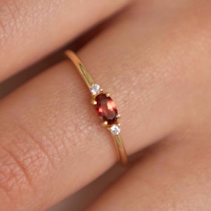 May include: A gold ring with a red oval gemstone and two smaller clear gemstones.