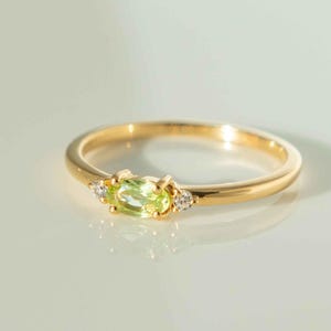 May include: A gold ring featuring a rectangular, light green gemstone, flanked by two small, round, clear gemstones. The ring has a thin band and is set against a soft, white background. This elegant ring is a piece of jewelry.
