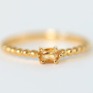 May include: A gold ring with a small, rectangular, light orange gemstone at the center. The band is adorned with small, evenly spaced gold beads. The ring is set against a white background.