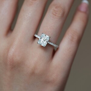 May include: A radiant-cut diamond engagement ring with a pavé band. The ring features a large, rectangular diamond set in a delicate, sparkling band. The ring is on a person's finger, with a blurred background.