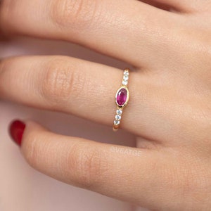 May include: A delicate gold ring with a single oval-shaped ruby gemstone surrounded by smaller white gemstones.