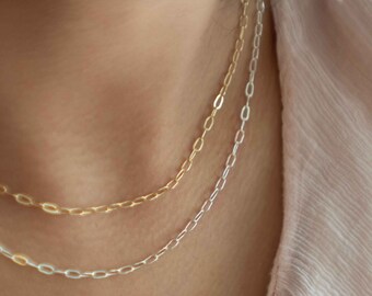 May include: Two delicate necklaces, one gold and one silver, rest against a person's skin. The necklaces feature a chain link design. The person is wearing a light pink, textured top.