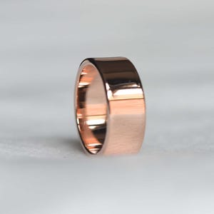 May include: A rose gold ring with a flat, wide band. The ring has a polished, reflective surface, catching the light. The ring is a simple, elegant piece of jewelry.