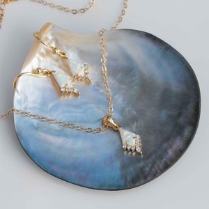 May include: A gold necklace and earrings set with a white opal and clear crystal accents. The jewelry is displayed on a mother-of-pearl shell dish. The brand name "VEATGE" is printed below the dish.