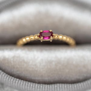 May include: A gold ring with a small, oval-shaped, bright pink gemstone. The band is adorned with small, round beads. The ring is presented in a gray velvet ring box.