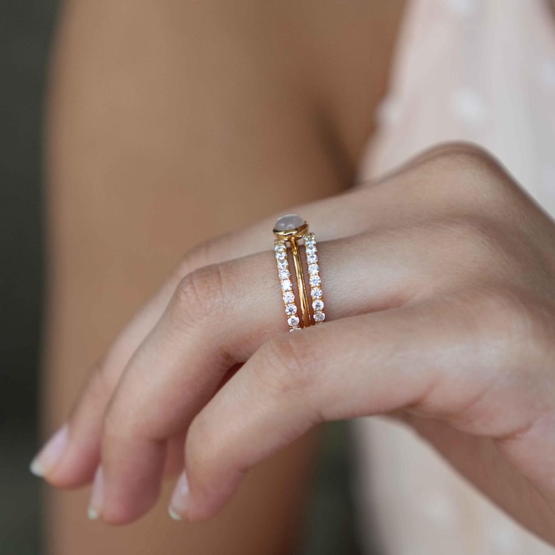 Pu&ograve; includere: Un set di tre anelli in oro indossati su un dito. L'anello centrale presenta una pietra cabochon rotonda e liscia di colore grigio pallido in una montatura a castone. &Egrave; affiancato da due fasce sottili e delicate, ciascuna con una fila di piccole pietre bianche.