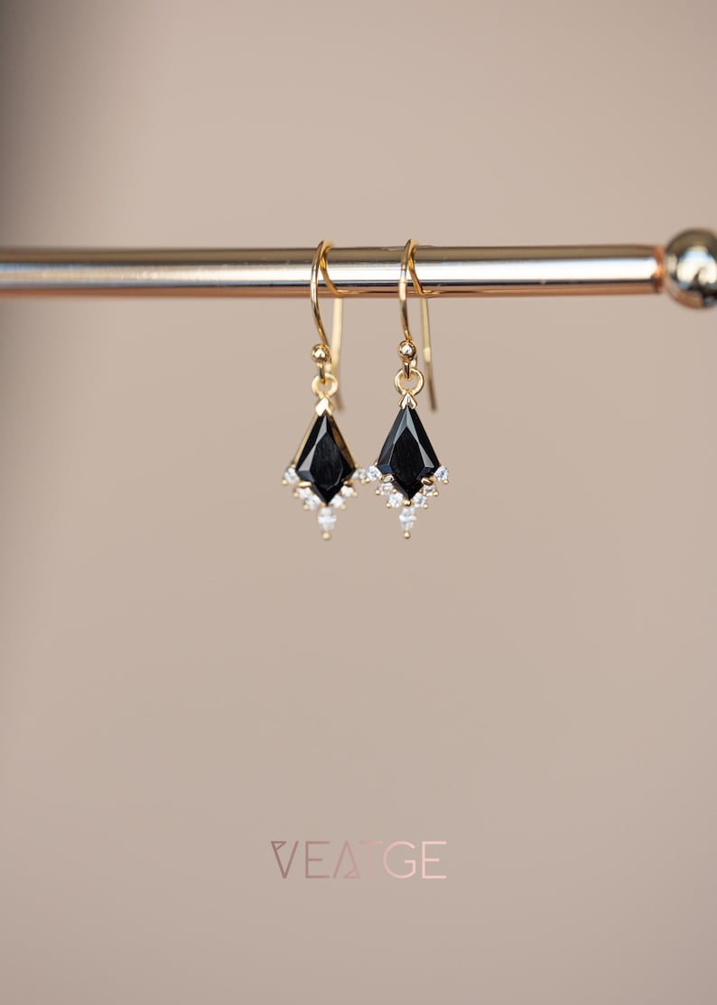 Peut inclure: Une paire de boucles d'oreilles pendantes dor&eacute;es. Chaque boucle d'oreille pr&eacute;sente une pierre noire en forme de losange avec un groupe de petites pierres blanches en bas. Les boucles d'oreilles sont suspendues &agrave; un crochet dor&eacute;.
