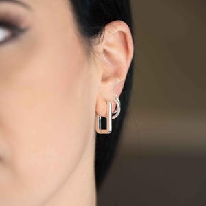 May include: A pair of silver-toned earrings consisting of a rectangular hoop earring worn in the lower lobe and a smaller circular hoop earring worn in the upper lobe. The earrings feature a polished metallic finish and a simple hinged design.