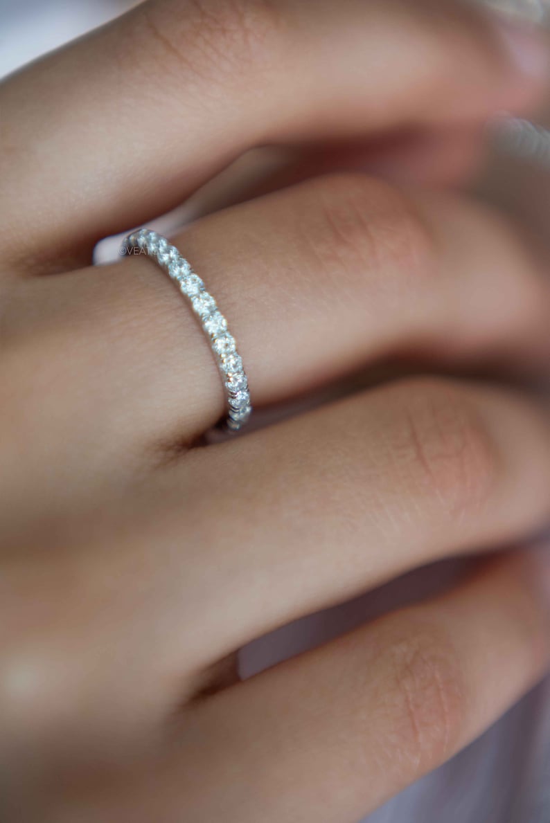 Pu&ograve; includere: Un anello a fascia in tonalit&agrave; argento con una fila continua di piccole pietre rotonde trasparenti sfaccettate, incastonate in un binario di metallo lucido. L'anello &egrave; indossato su un dito, evidenziando la brillantezza delle pietre.