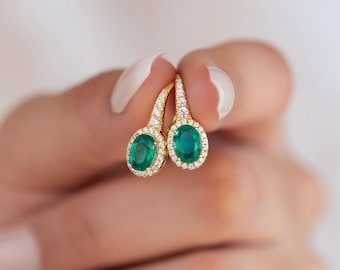 May include: A pair of gold earrings featuring oval emerald-green gemstones. Each earring is encircled by a halo of small, clear gemstones. The earrings are held up by a person's fingers.