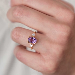 May include: A gold ring featuring an oval-cut purple gemstone, flanked by two pear-shaped white gemstones. The ring is set on a slender gold band. The ring is displayed on a person's finger.