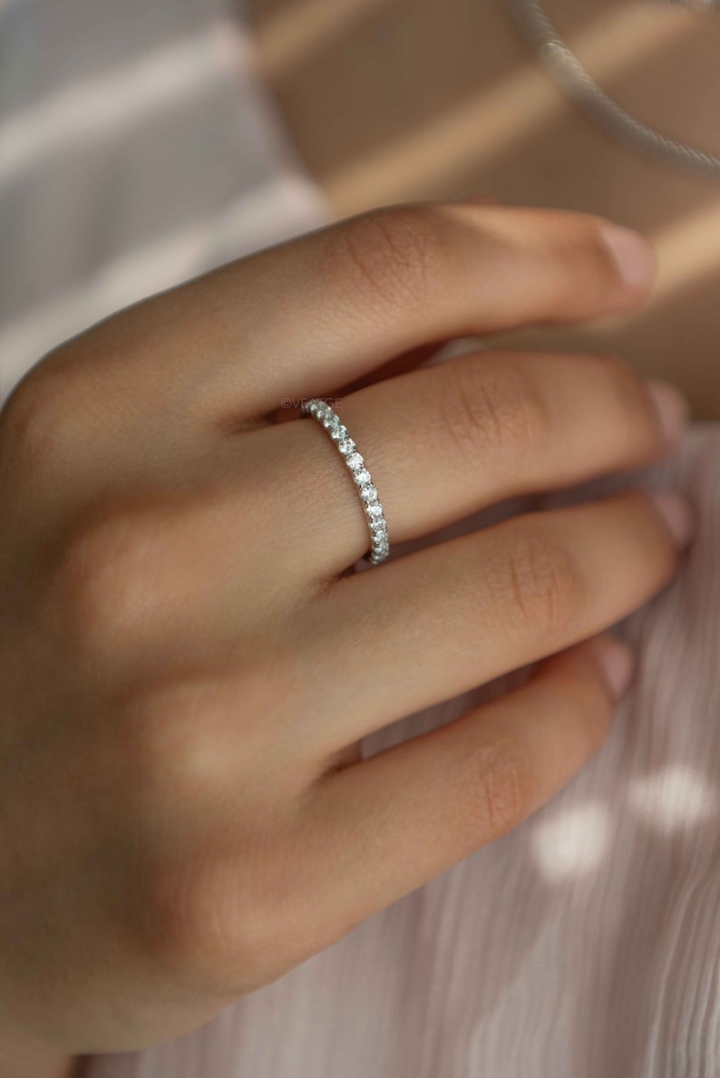 Pu&ograve; includere: Un sottile anello color argento caratterizzato da una fila continua di piccole pietre chiare a taglio rotondo lungo l'intera circonferenza esterna. La fascia in metallo ha una finitura lucida e le pietre sono disposte in modo uniforme.