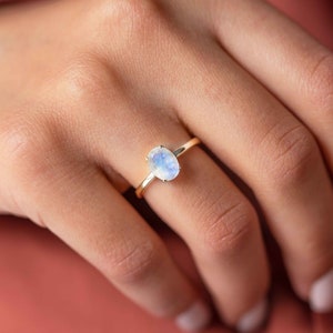 May include: A gold ring featuring an oval, iridescent moonstone. The gemstone is set on a thin, yellow gold band. The ring is displayed on a person's hand.