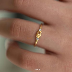 May include: A delicate gold ring featuring a central oval-cut, yellow gemstone, flanked by two smaller, round, clear gemstones. The ring is set on a thin gold band. The ring is shown on a person's finger.