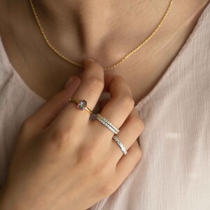 Pu&ograve; includere: Un anello in oro con una pietra ovale iridescente, accompagnato da due fasce color argento incastonate con piccole pietre chiare. &Egrave; visibile anche una sottile collana a catena dorata. I gioielli sono indossati sulle dita e al collo.