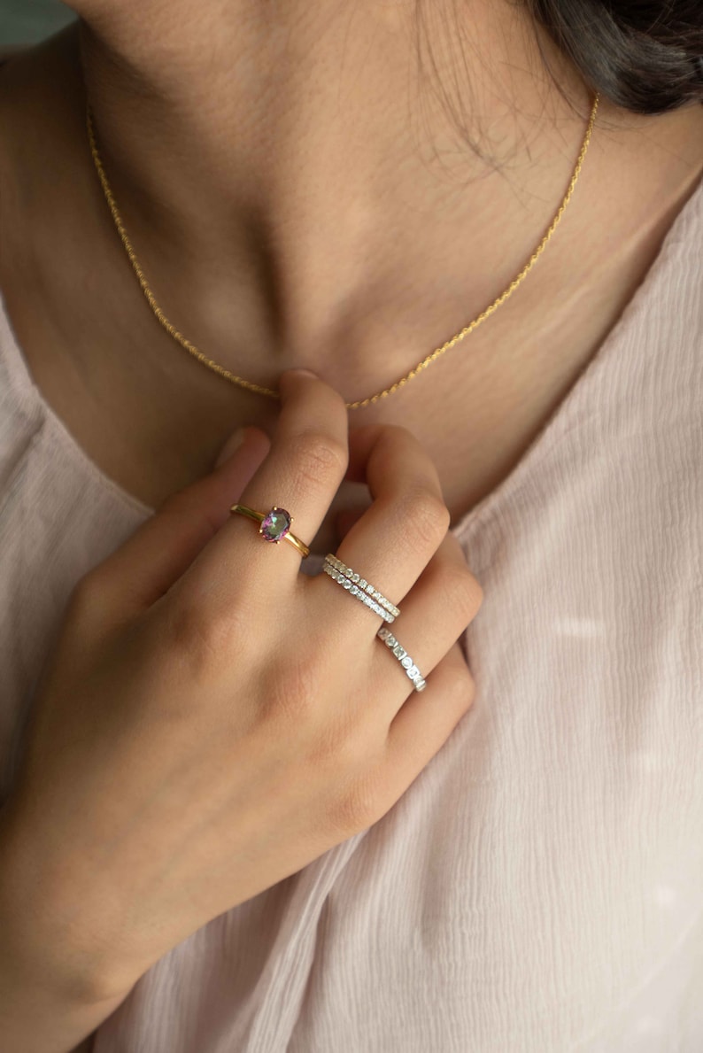 Pu&ograve; includere: Un anello in oro con una pietra ovale iridescente, accompagnato da due fasce color argento incastonate con piccole pietre chiare. &Egrave; visibile anche una sottile collana a catena dorata. I gioielli sono indossati sulle dita e al collo.