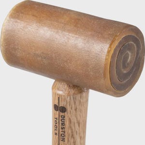May include: A rawhide mallet with a cylindrical head and a light-coloured wooden handle. The head has a smooth, tan, translucent surface with visible concentric layers. The handle features the black printed text DURSTON TOOLS with a logo.