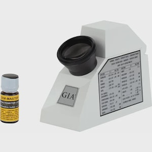 May include: A white GIA refractometer with a black eyepiece and a side-mounted refractive index reference chart, accompanied by a small brown glass bottle labeled Gem-Master Refractive Index Liquid with a white cap and black text.
