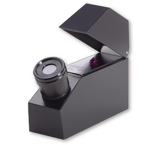 May include: A black metal refractometer featuring an angled eyepiece and a hinged lid. Inside the device, a small, faceted, deep purple gemstone is positioned on a reflective metal surface, visible through the open top of the instrument.