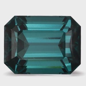 Può includere: Una gemma sciolta con taglio a smeraldo di colore blu-verde intenso. La pietra presenta una forma rettangolare con superfici sfaccettate che creano riflessi di luce geometrici sul corpo trasparente del cristallo blu-verde scuro.