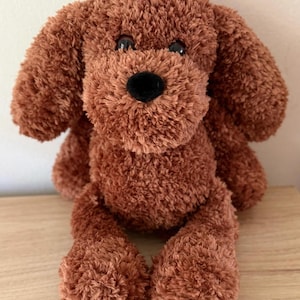 May include: A plush brown dog toy with floppy ears, a black nose, and black eyes. The stuffed animal is made of soft, textured material and is sitting on a wooden surface. The toy is designed to resemble a puppy.