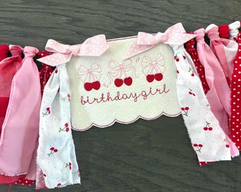May include: A fabric banner featuring a central off-white scalloped panel embroidered with three pairs of red cherries and the text birthday girl in red script. The panel is flanked by strips of red, pink, and white fabric with cherry patterns.