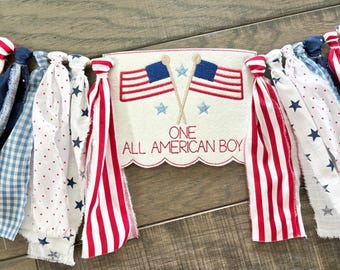 Puede incluir: Un estandarte de tela con un panel central blanco bordado con banderas estadounidenses cruzadas, estrellas azules y el texto rojo ONE ALL AMERICAN BOY. El estandarte está decorado con flecos de tiras de tela anudadas con patrones rojos, blancos y azules.