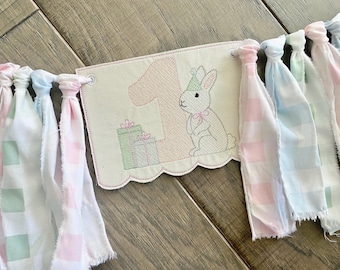 Puede incluir: Un estandarte de tela con un borde inferior festoneado y detalles bordados que incluyen el número uno, un conejo con un sombrero de fiesta y dos cajas de regalo. El estandarte está decorado con tiras anudadas de tela a cuadros en tonos pastel.
