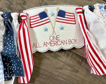 May include: A fabric banner featuring a scalloped white felt center panel embroidered with two crossed American flags, three light blue stars, and the red text ONE ALL AMERICAN BOY. The banner is accented with tied strips of red, white, and blue fabric.