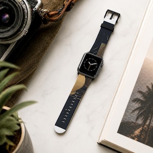 May include: Black smartwatch with a patterned band featuring gold and black geometric designs. The watch band has a white tip and a black buckle. The watch is displayed on a white surface next to a camera and a book.