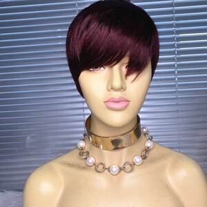 May include: A mannequin head with a short, burgundy-colored wig. The wig has a side-swept bang. The mannequin is wearing a gold-colored choker necklace and a pearl necklace. The background is a window with horizontal blinds.