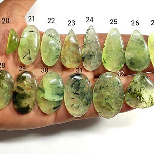 May include: A selection of polished prehnite gemstones in diverse shapes, such as oval, teardrop, and triangular. The stones vary in colour from pale to vibrant green, with dark inclusions. The gemstones are shown against a light background.