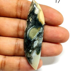 May include: An elongated, marquise-shaped polished gemstone cabochon featuring a marbled pattern of cream, dark charcoal, and sage green colors. The stone has a smooth, glossy surface with natural inclusions and the number 17 printed in black.